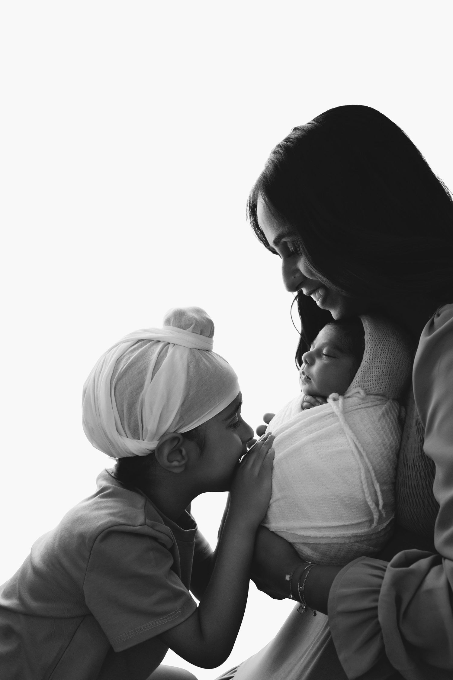 Black and white photo of a mother holding a newborn while a young child in a turban lovingly kisses the baby.
