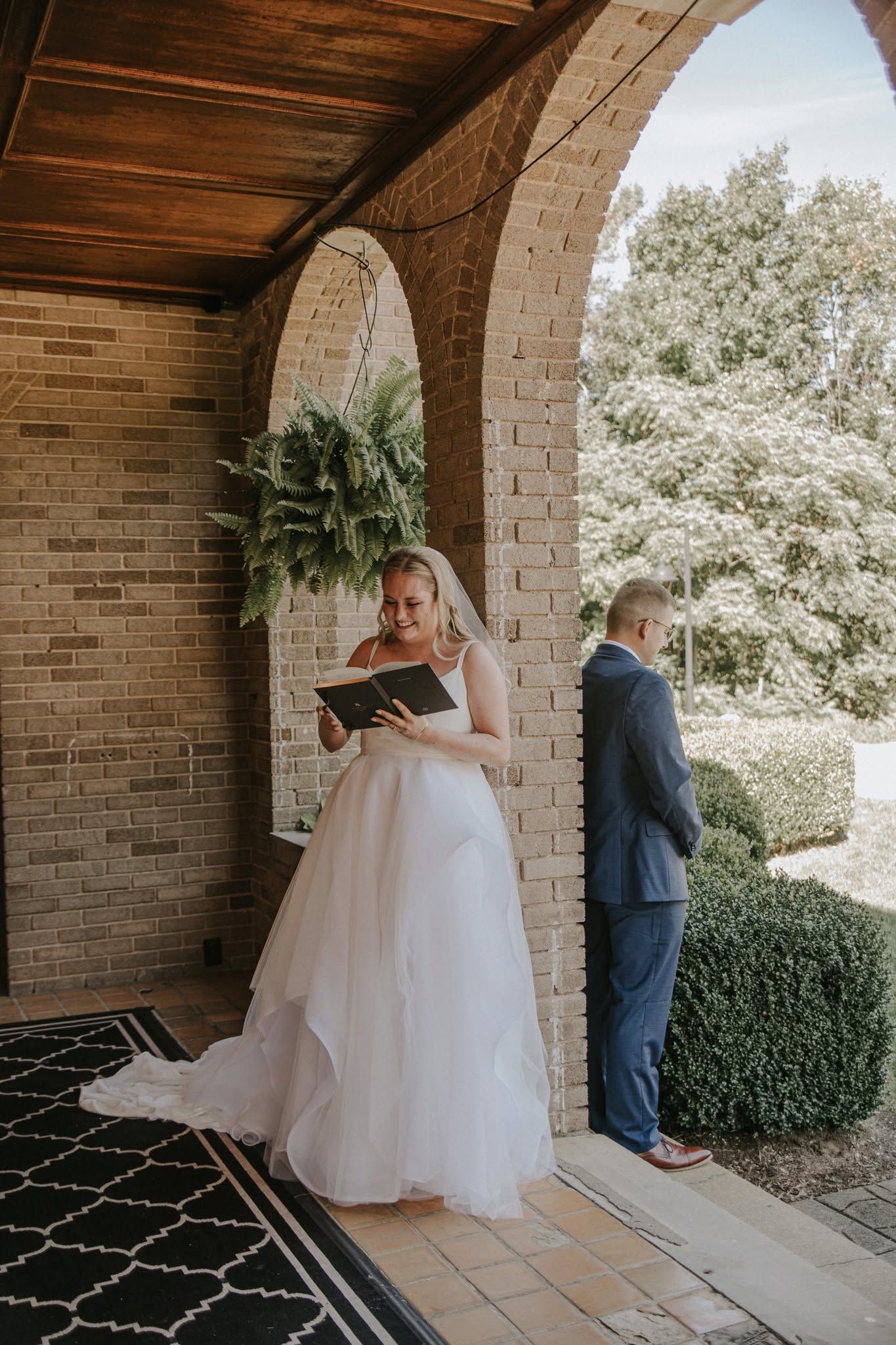 Bride in white gown reads vows from black book while groom stands facing brick arch at outdoor wedding ceremony.