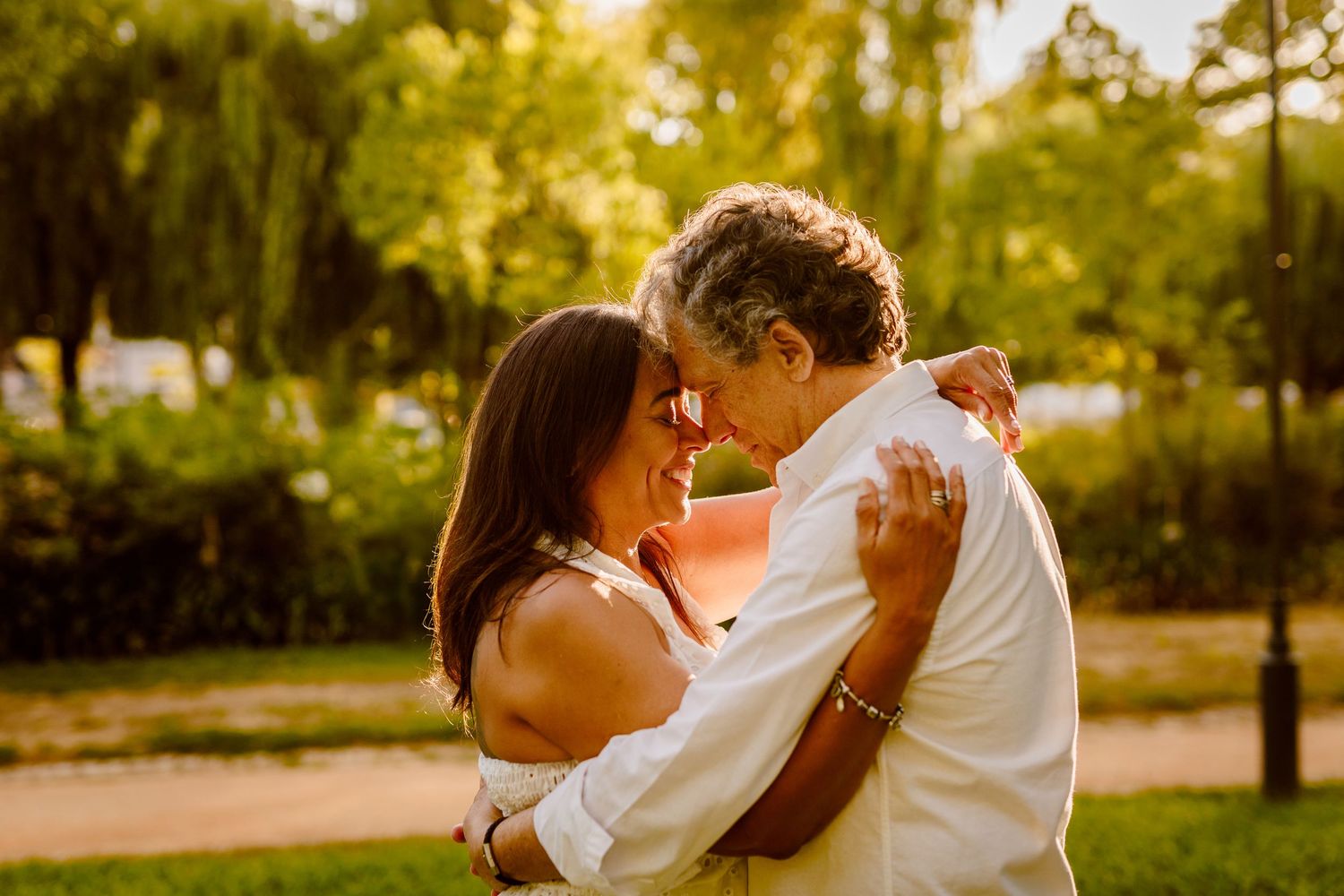Couple sitting together by water, woman with tattoos in white dress and pearl earrings, man in dark jacket, golden hour light.
