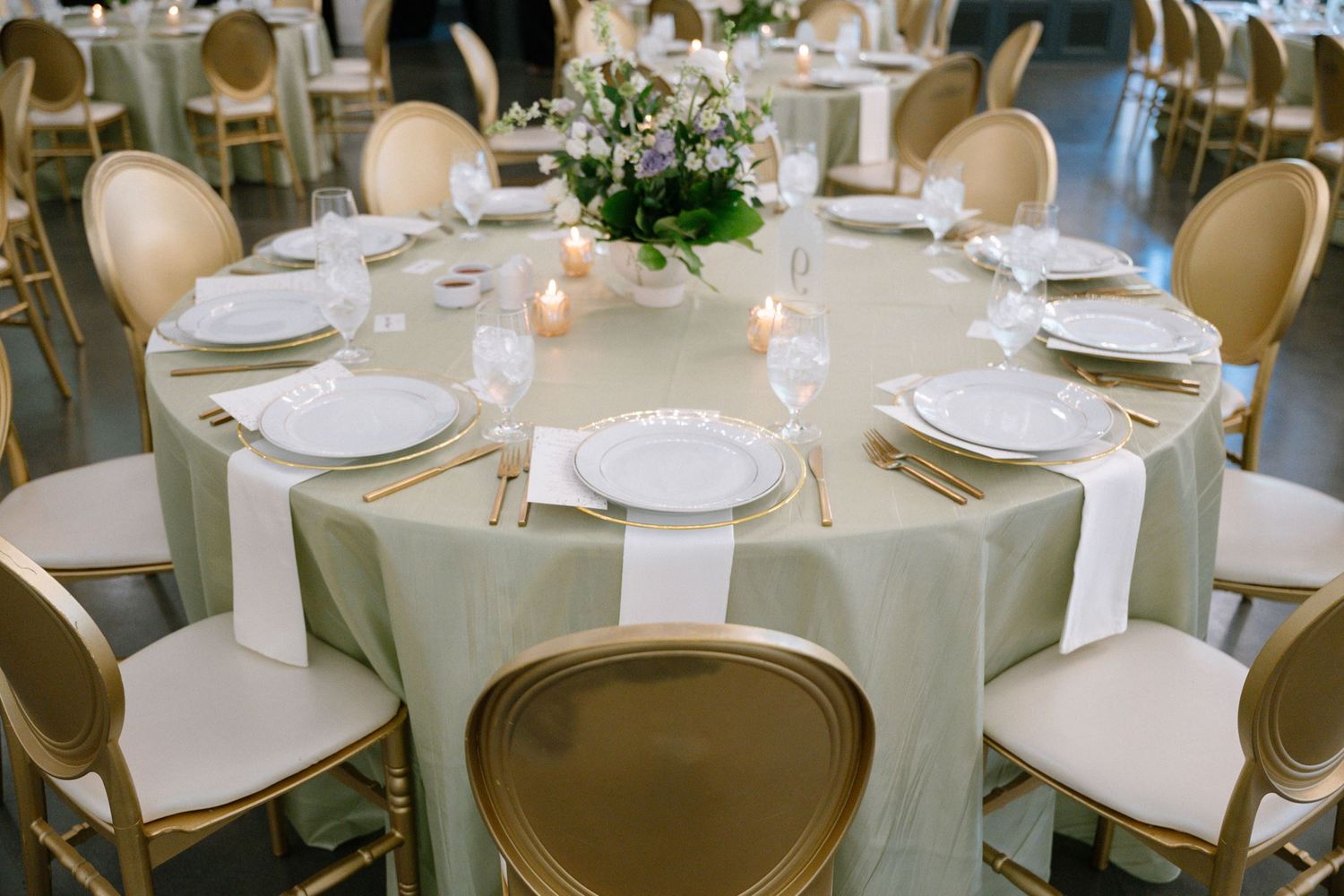 Elegant round wedding reception table with sage green linen, gold chairs, white plates, candles, and floral centerpiece.