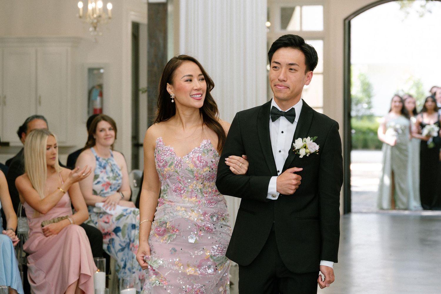 A smiling couple walks arm-in-arm at a wedding, man in tuxedo with boutonniere, woman in floral pink gown.