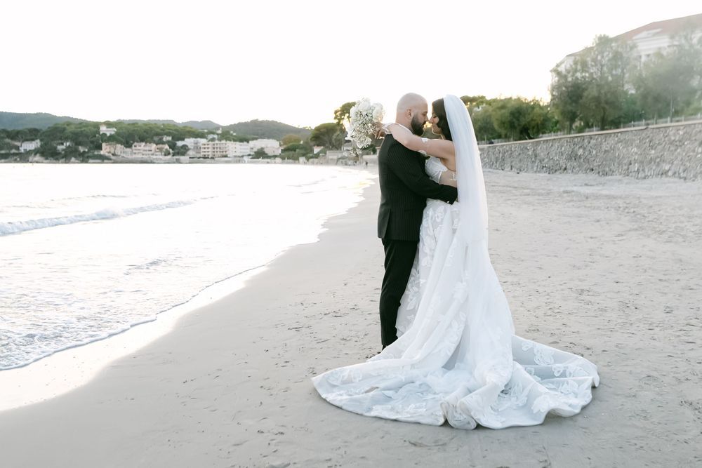 Bride and groom share a romantic kiss on a sandy beach, her long white lace veil flowing behind them.