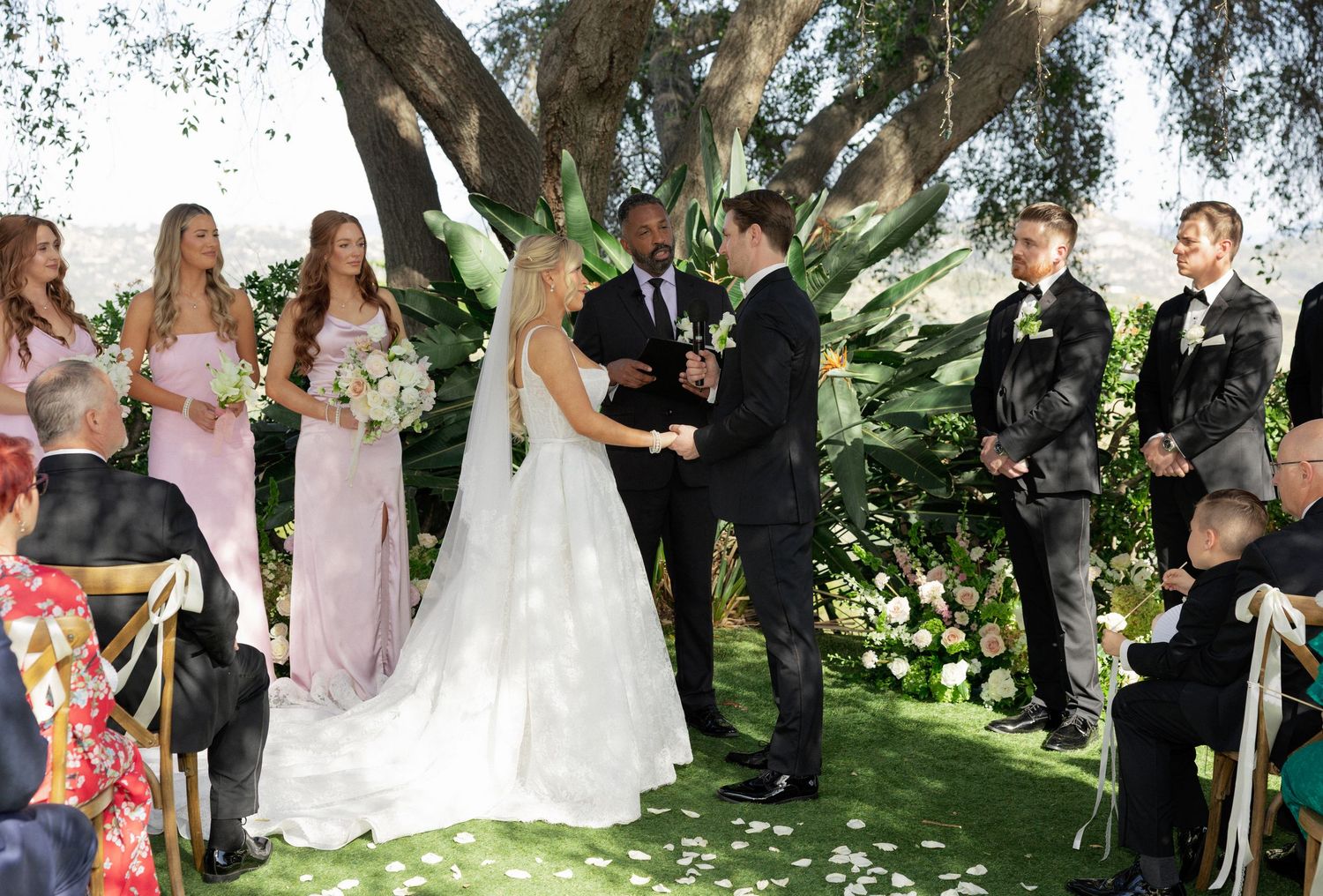 Outdoor wedding ceremony under a large oak tree with bride and groom at the altar surrounded by guests seated on white chairs.