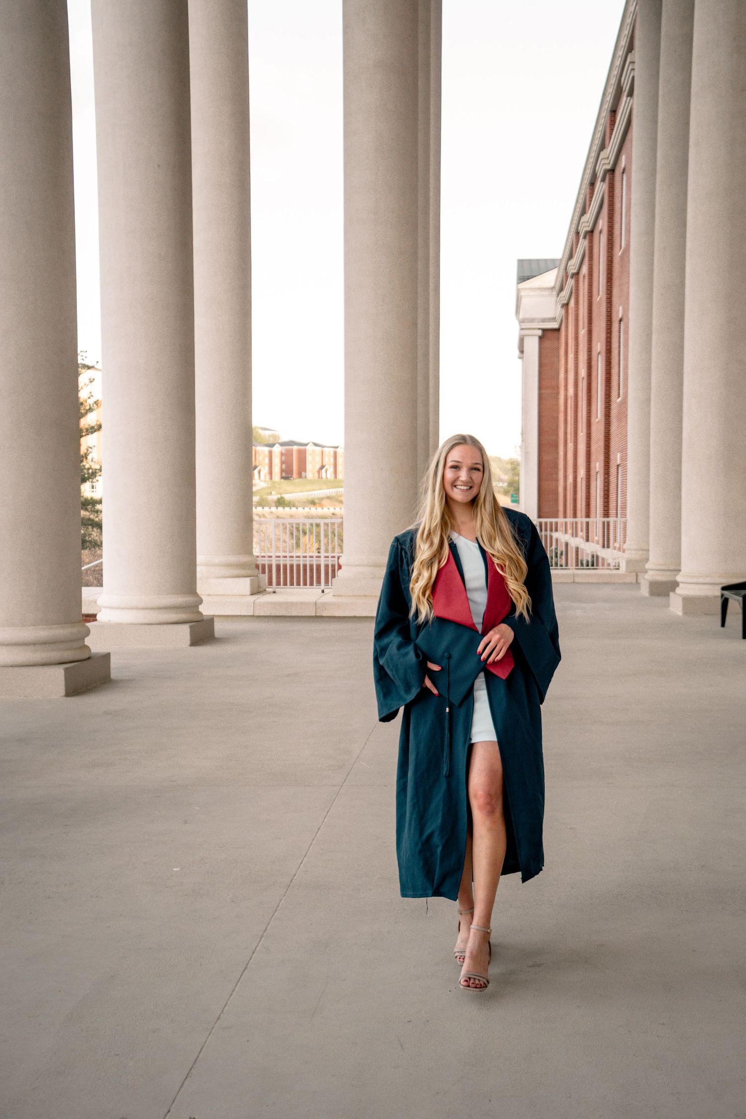 Smiling female graduate in dark cap and gown with red stole poses between stone columns in white dress.