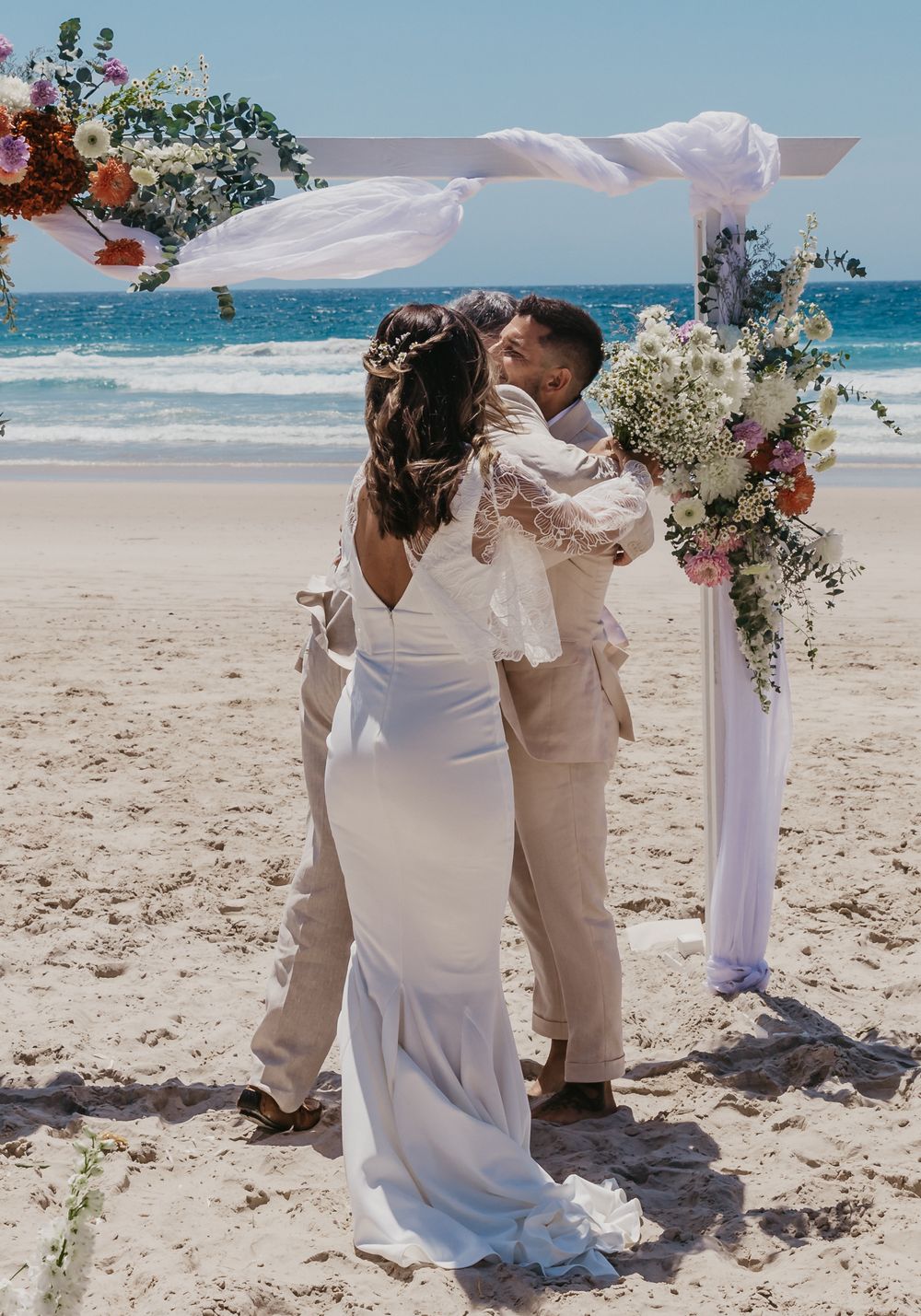 Bride and groom sharing a joyful embrace beneath a floral arbour after saying "I do" on Byron Bay beach.
