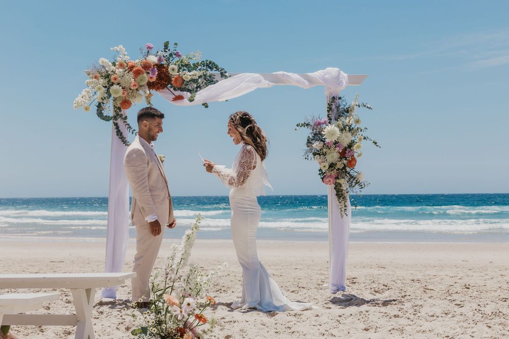 Bride and groom exchanging vows beneath a floral arbour during their intimate Byron Bay beach wedding ceremony.