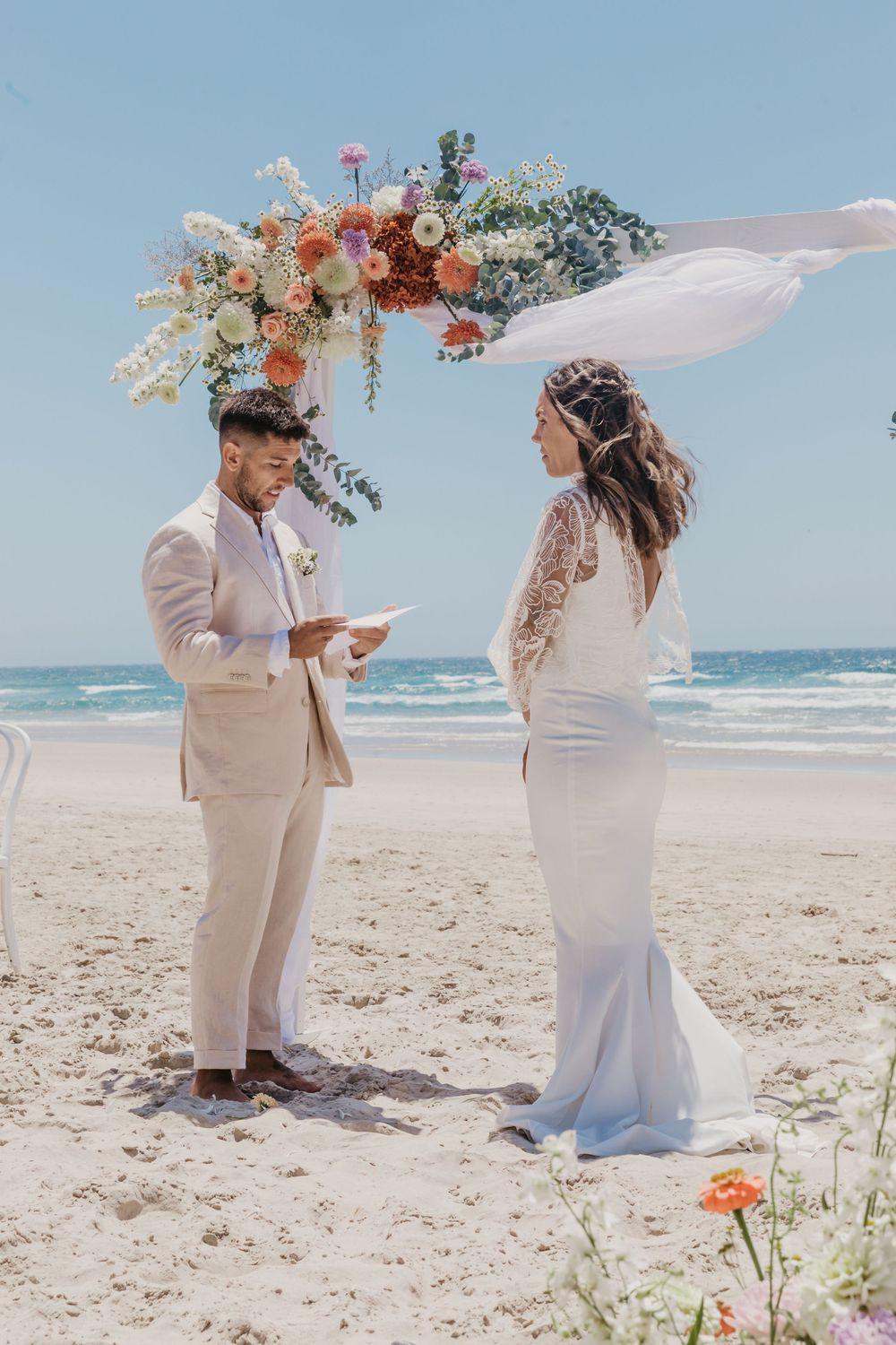 Bride and groom exchanging heartfelt vows beneath a floral arbour on the sands of Byron Bay beach.
