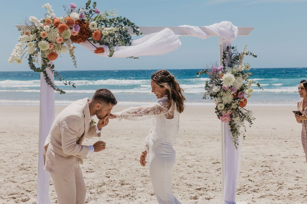 Groom kissing the bride's hand beneath a floral arbour after their romantic Byron Bay beach wedding ceremony.