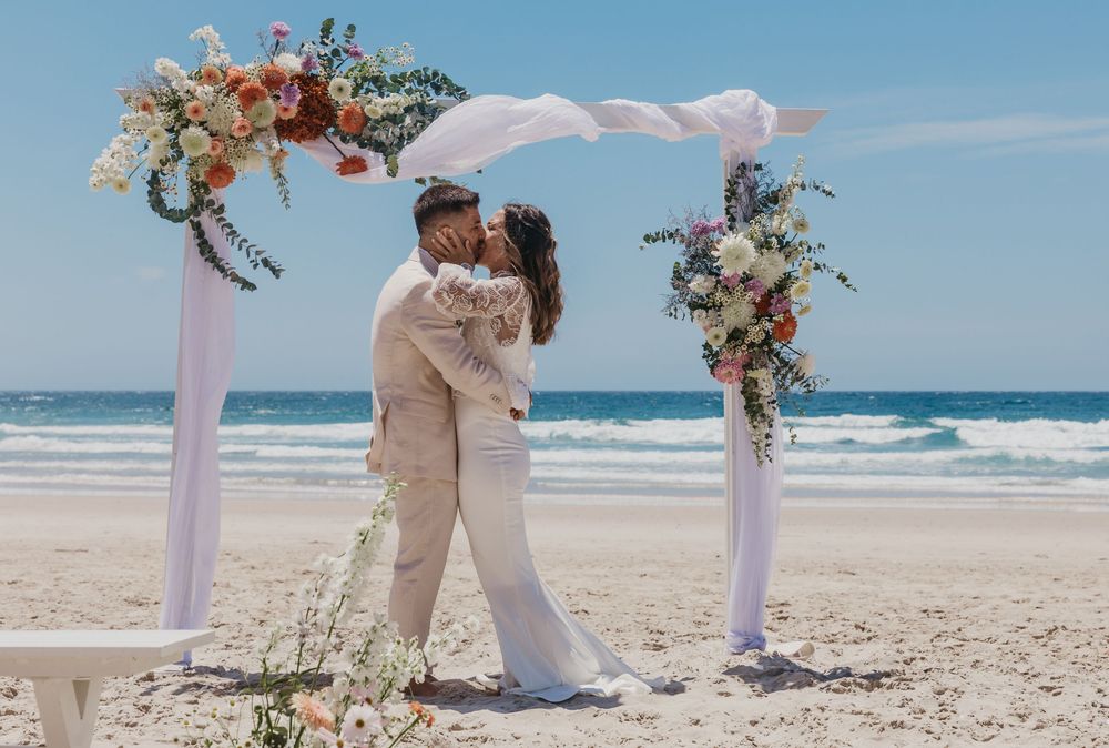 Bride and groom sharing their first kiss beneath a floral arbour during an intimate Byron Bay beach wedding ceremony.