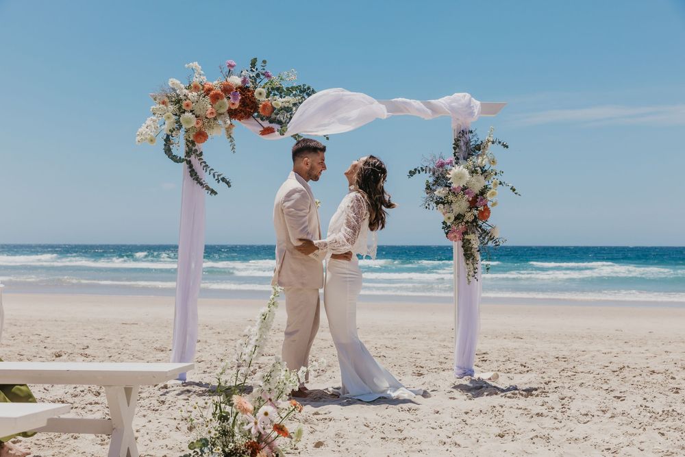 Bride and groom exchanging vows beneath a floral arbour during a romantic Byron Bay beach wedding ceremony.