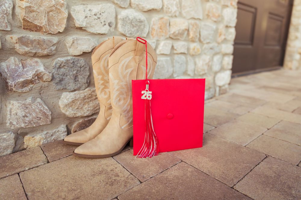 Tan cowboy boots beside a red graduation cap and diploma on a stone patio near a brick wall.