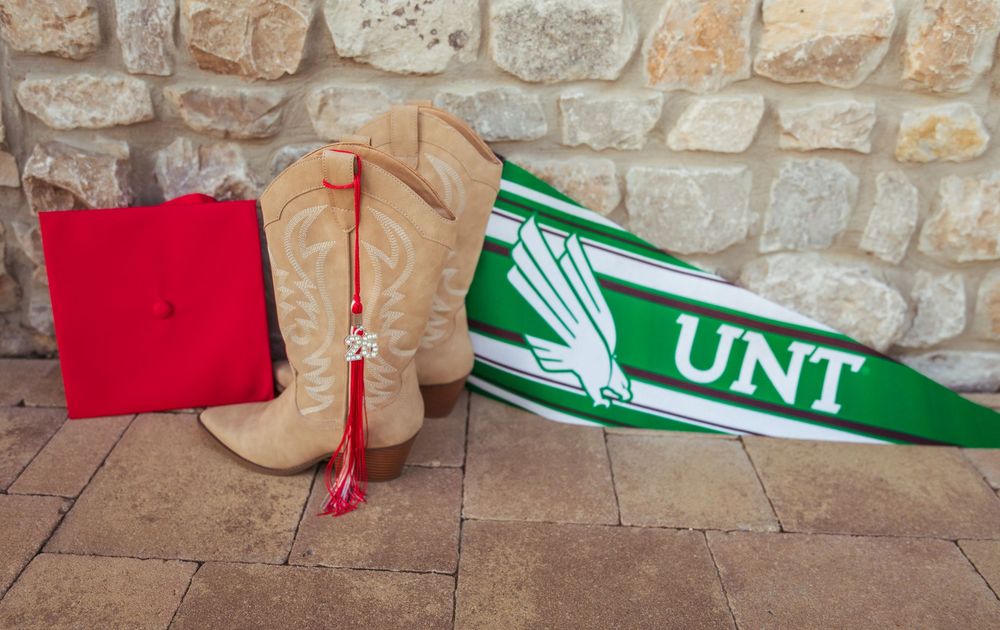 UNT green pennant with cowboy boots, red graduation cap with tassel, University of North Texas grad photo.