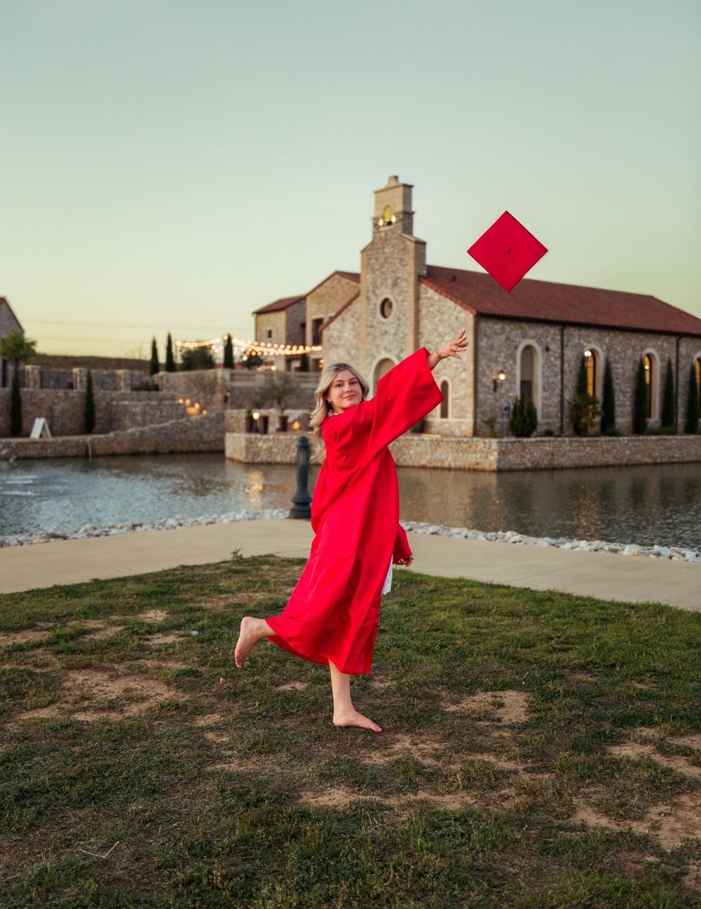 A joyful graduate in red cap and gown tosses her cap near a scenic stone chapel at sunset.