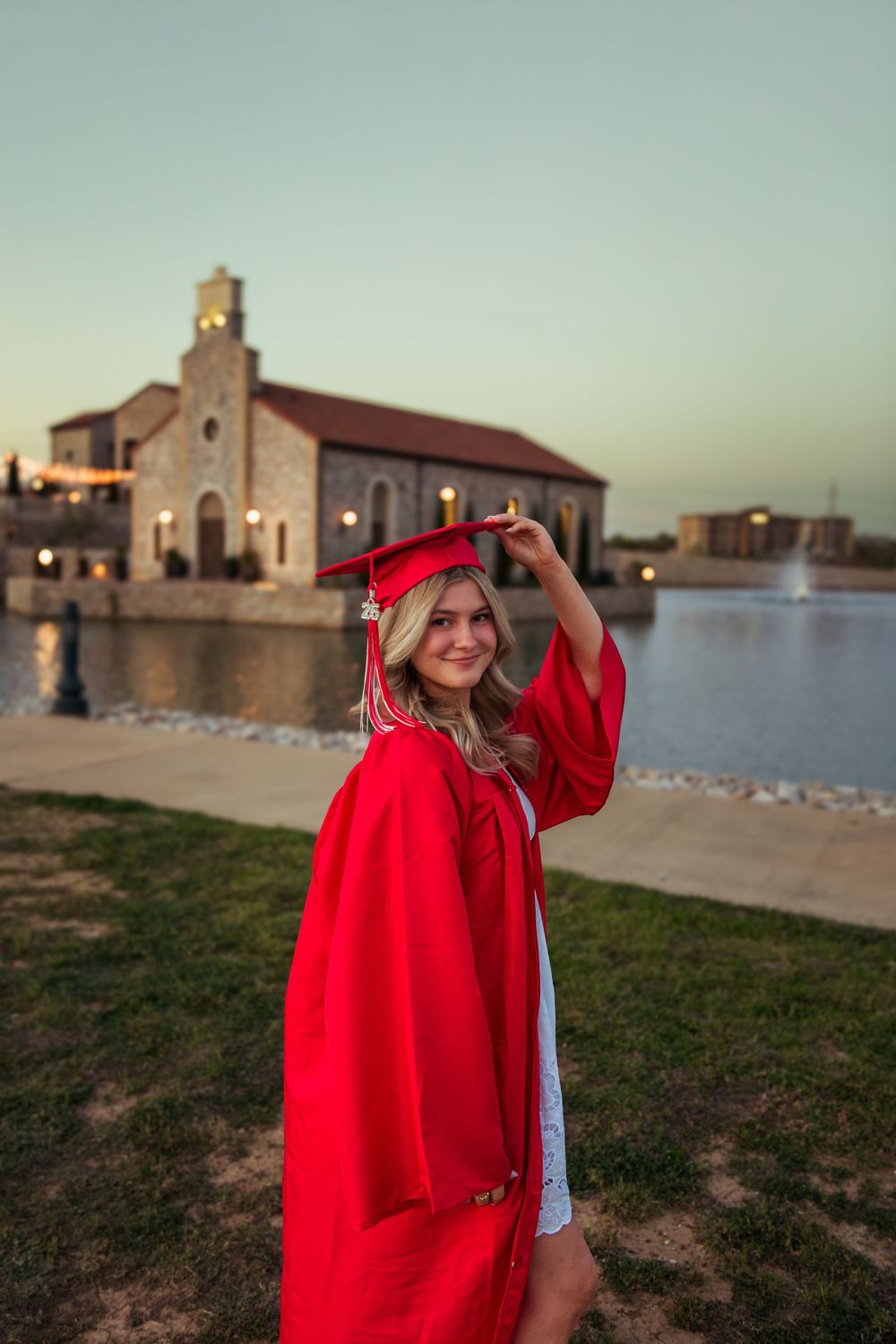 Smiling young woman in red graduation cap and gown posing outdoors near a lake and historic building at dusk.