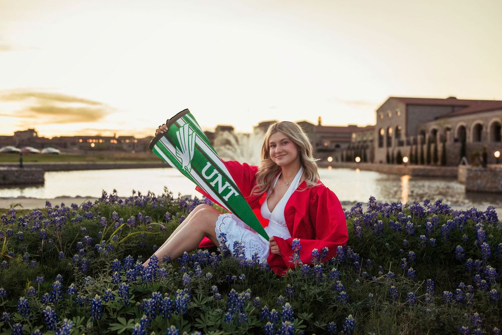 Woman in red jacket holding UNT pennant sits among bluebonnets at sunset near a lake.