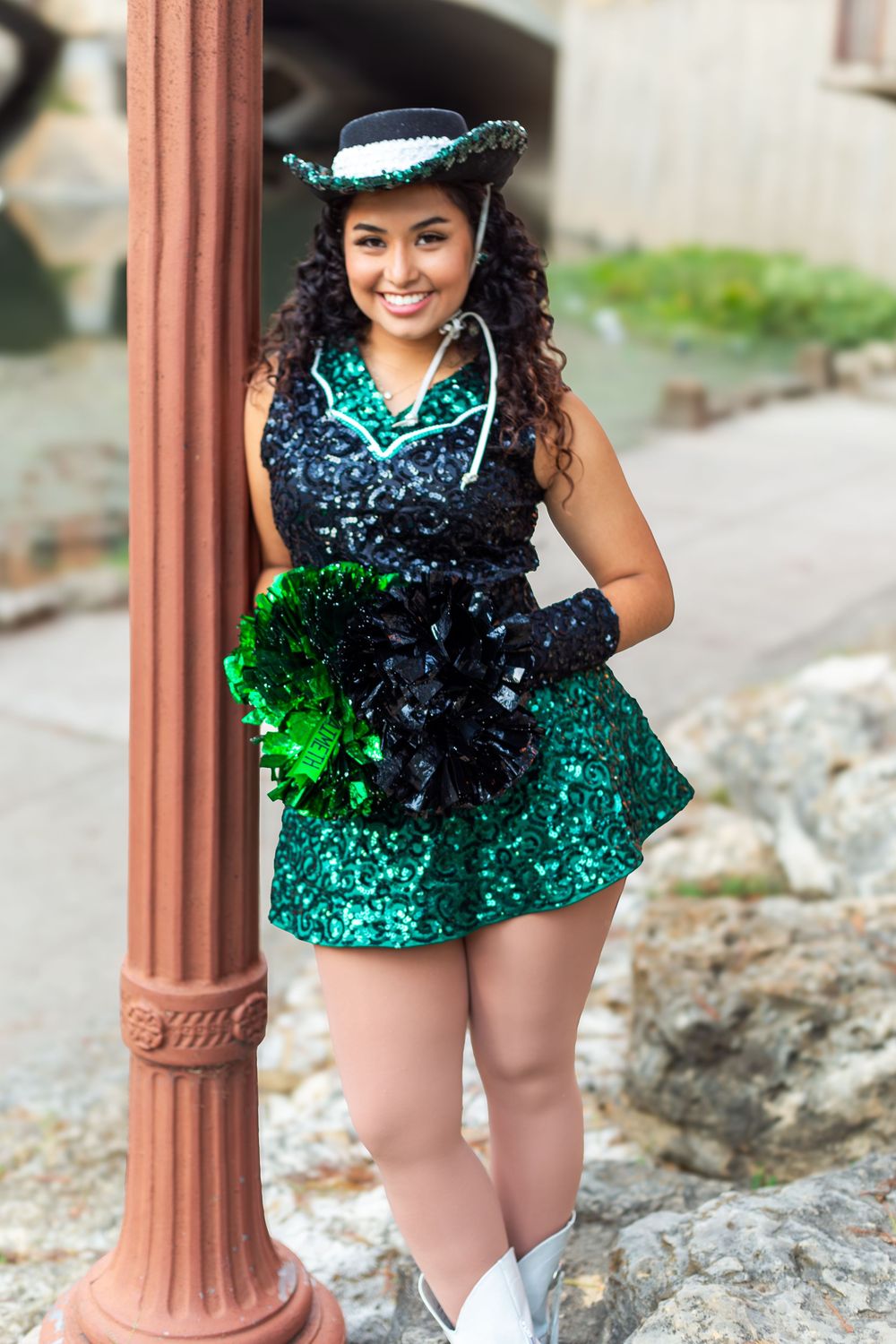 Young woman in green sequin cheerleader outfit with black pom-poms and hat, smiling beside a lamppost.