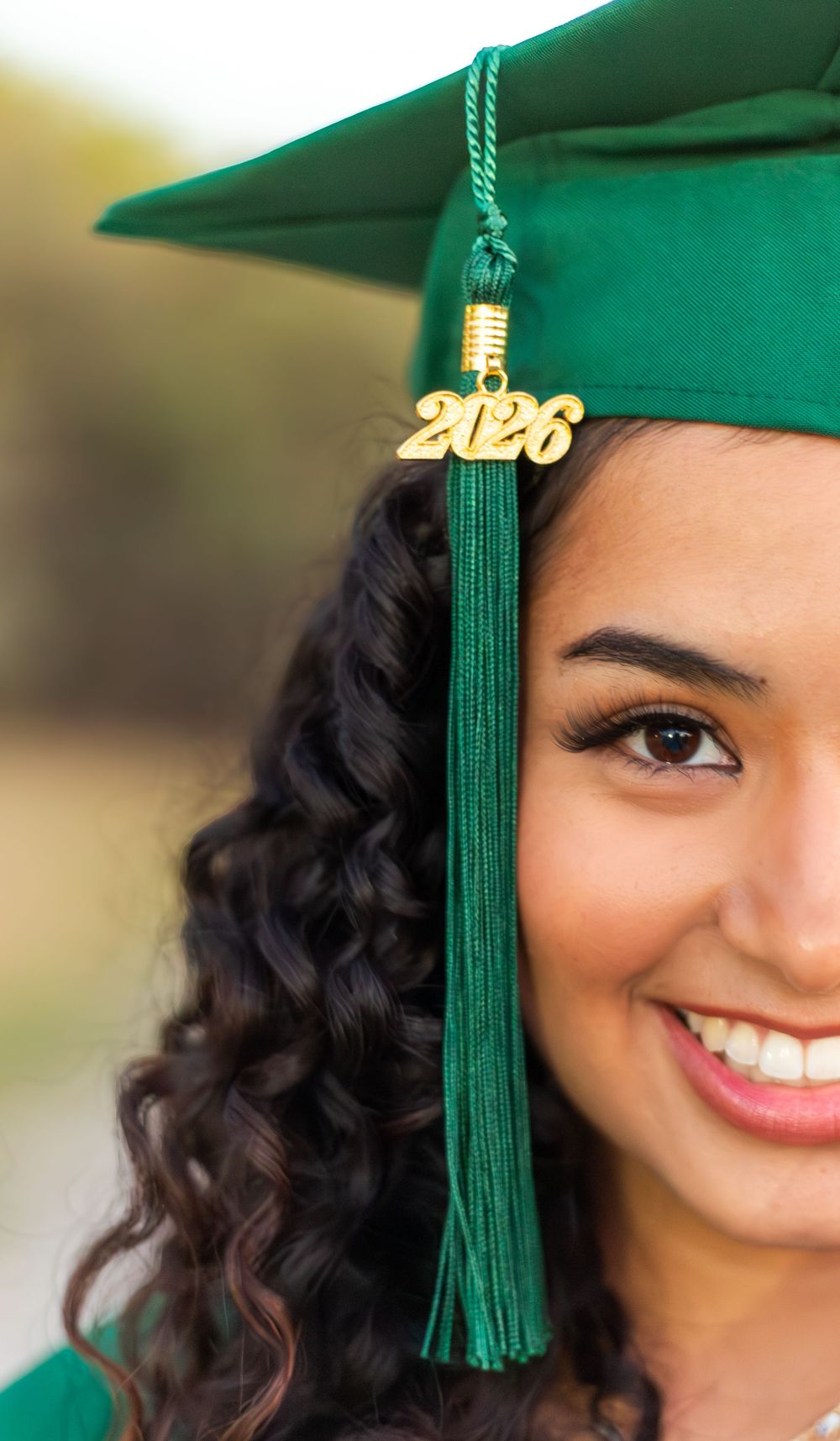 Smiling young woman in green graduation cap and gown with gold 2026 tassel charm celebrating her achievement.