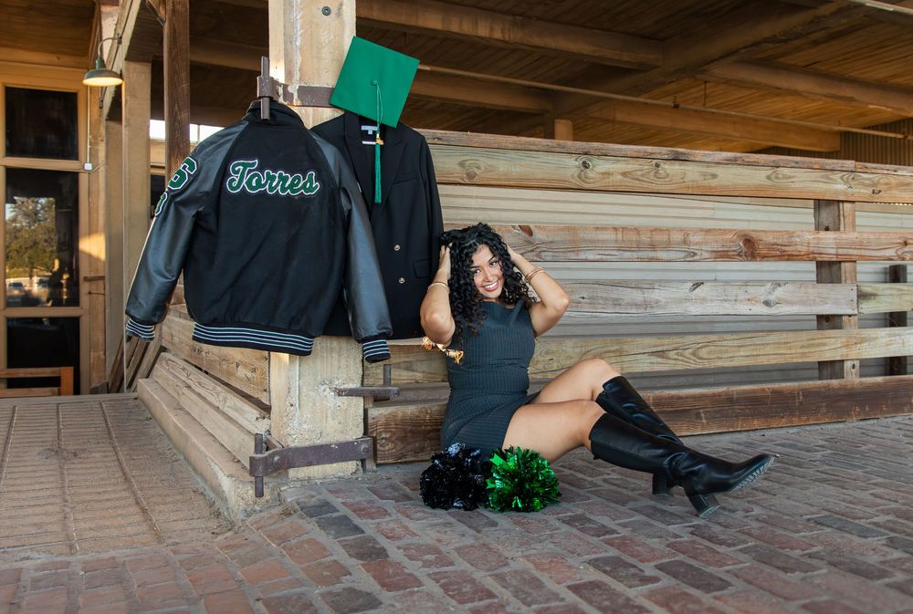 Woman in black dress and boots poses beside a varsity jacket display on a brick patio.