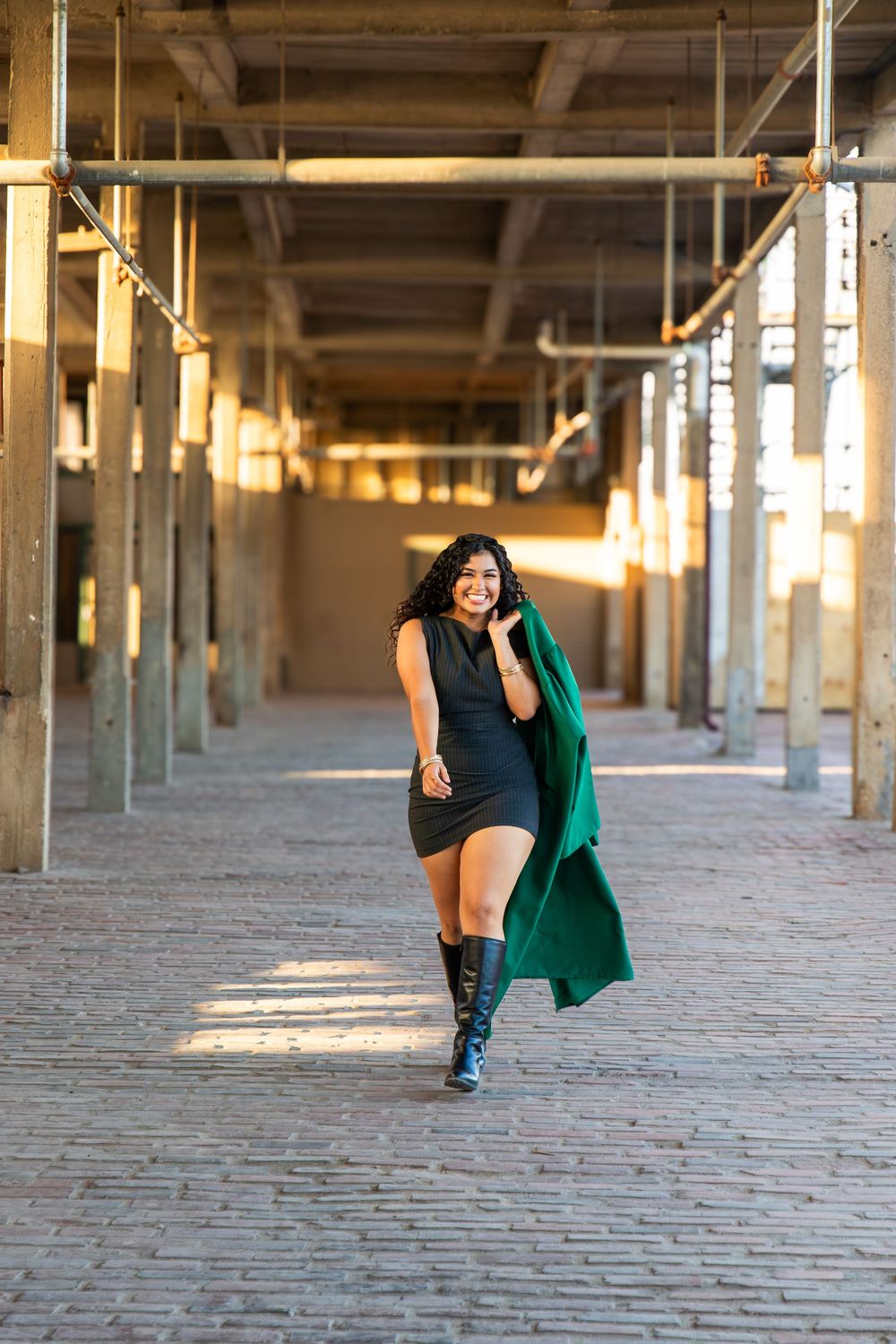 Woman in black dress and green coat walks confidently through an industrial corridor with brick floors and golden sunlight.