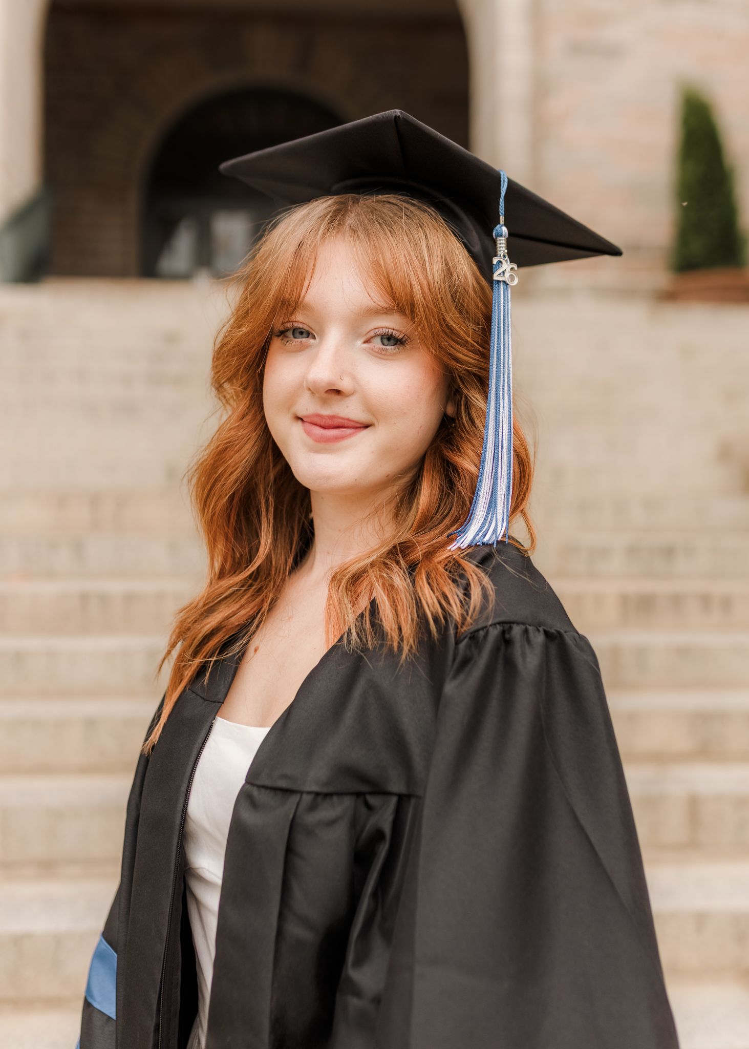 Young woman with red hair smiling in black graduation cap and gown, standing on university steps.