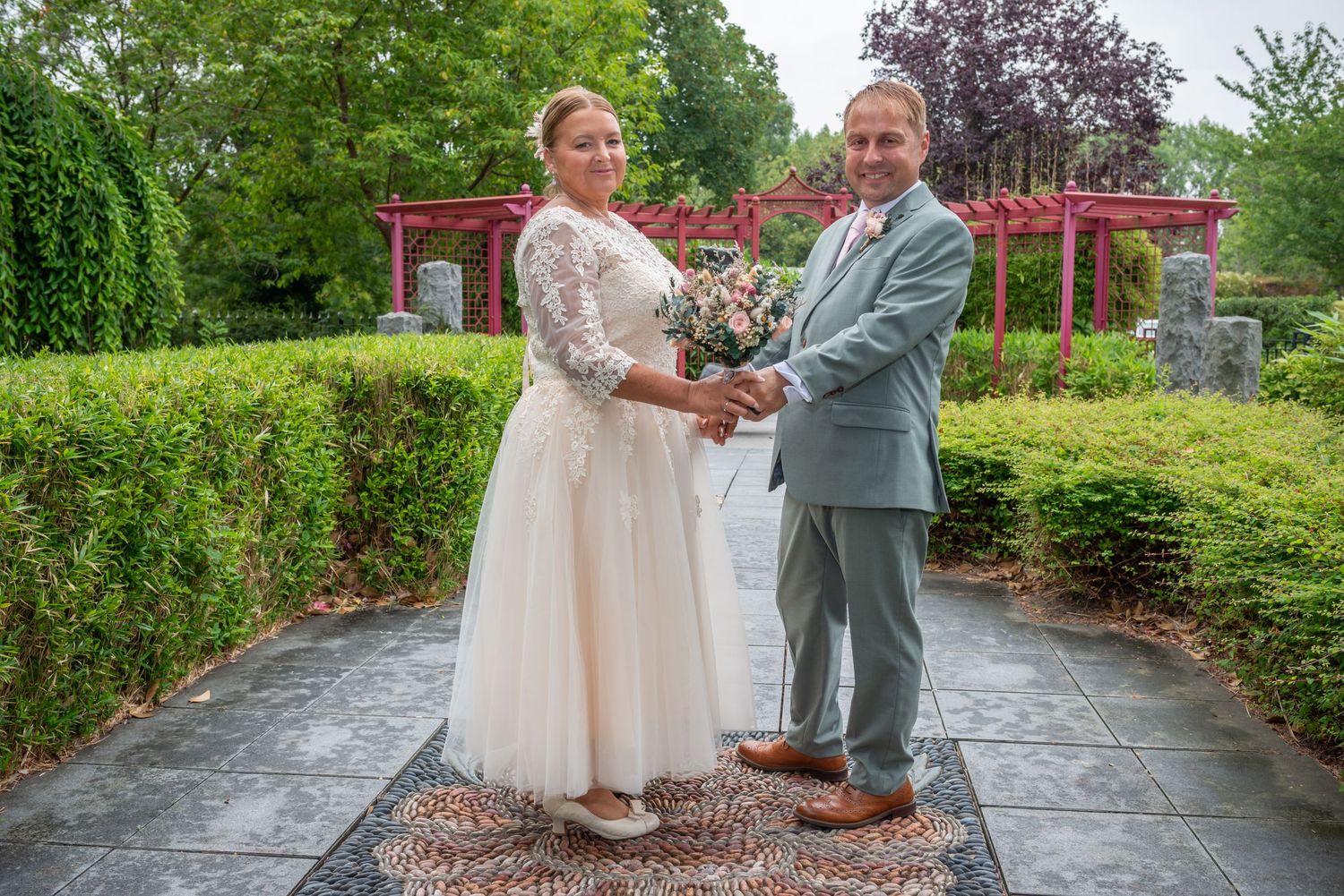 A smiling couple in wedding attire holds hands in a lush garden, bride in white lace gown, groom in gray suit.