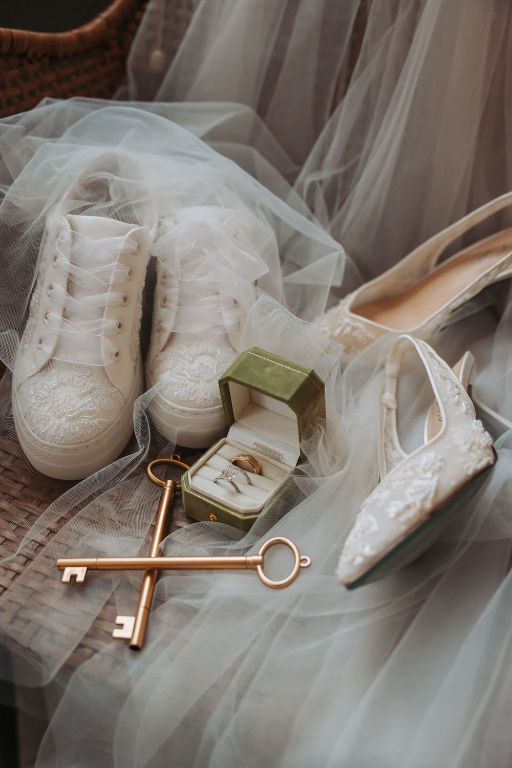 Bridal flat lay with lace heels, white sneakers, gold keys, and a green velvet ring box on sheer tulle fabric.