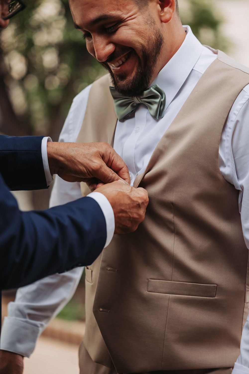 Smiling groom in tan vest and green bow tie gets boutonniere pinned by groomsman outdoors.