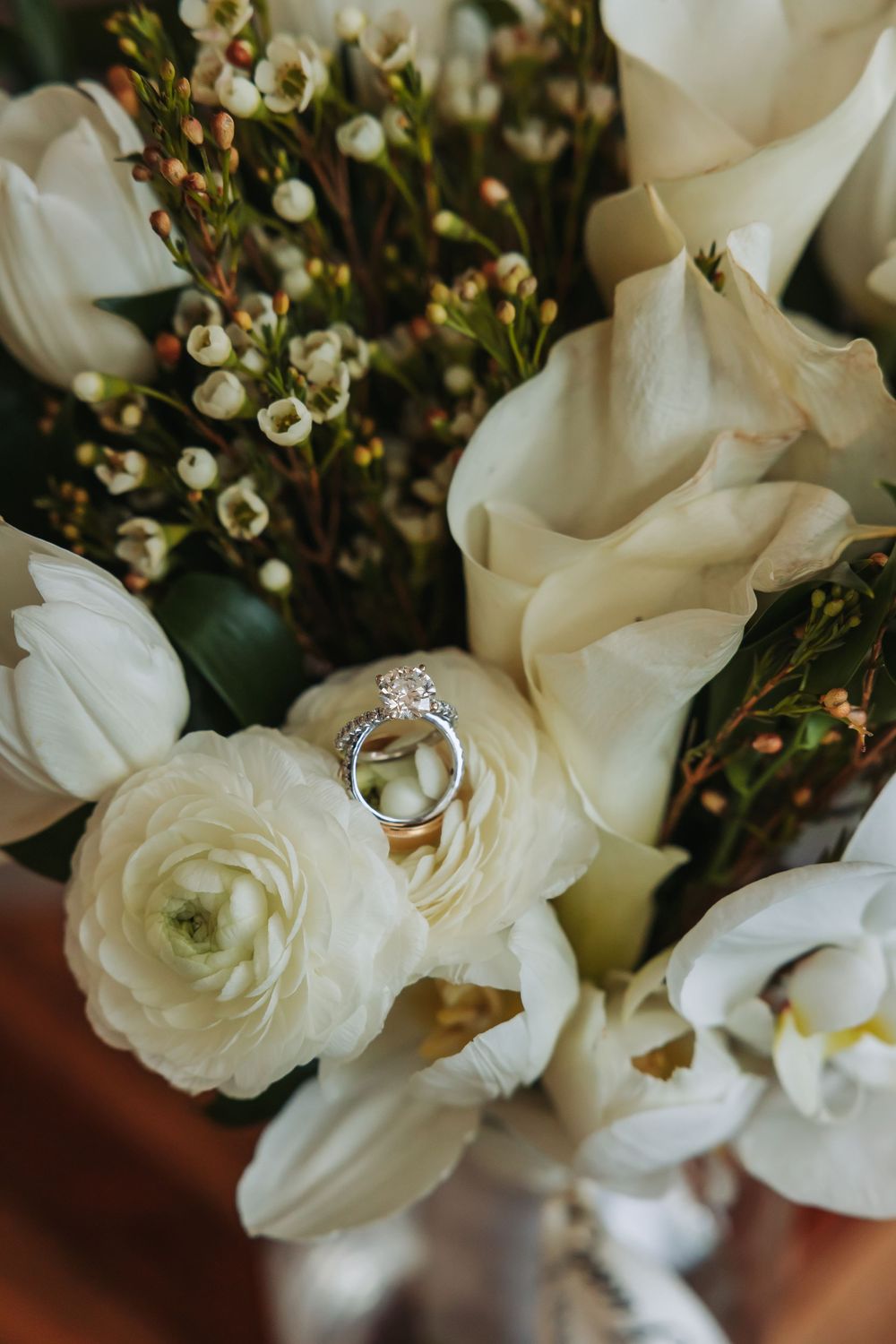 Wedding rings nestled among white ranunculus, calla lilies, and wax flowers in an elegant bridal bouquet.