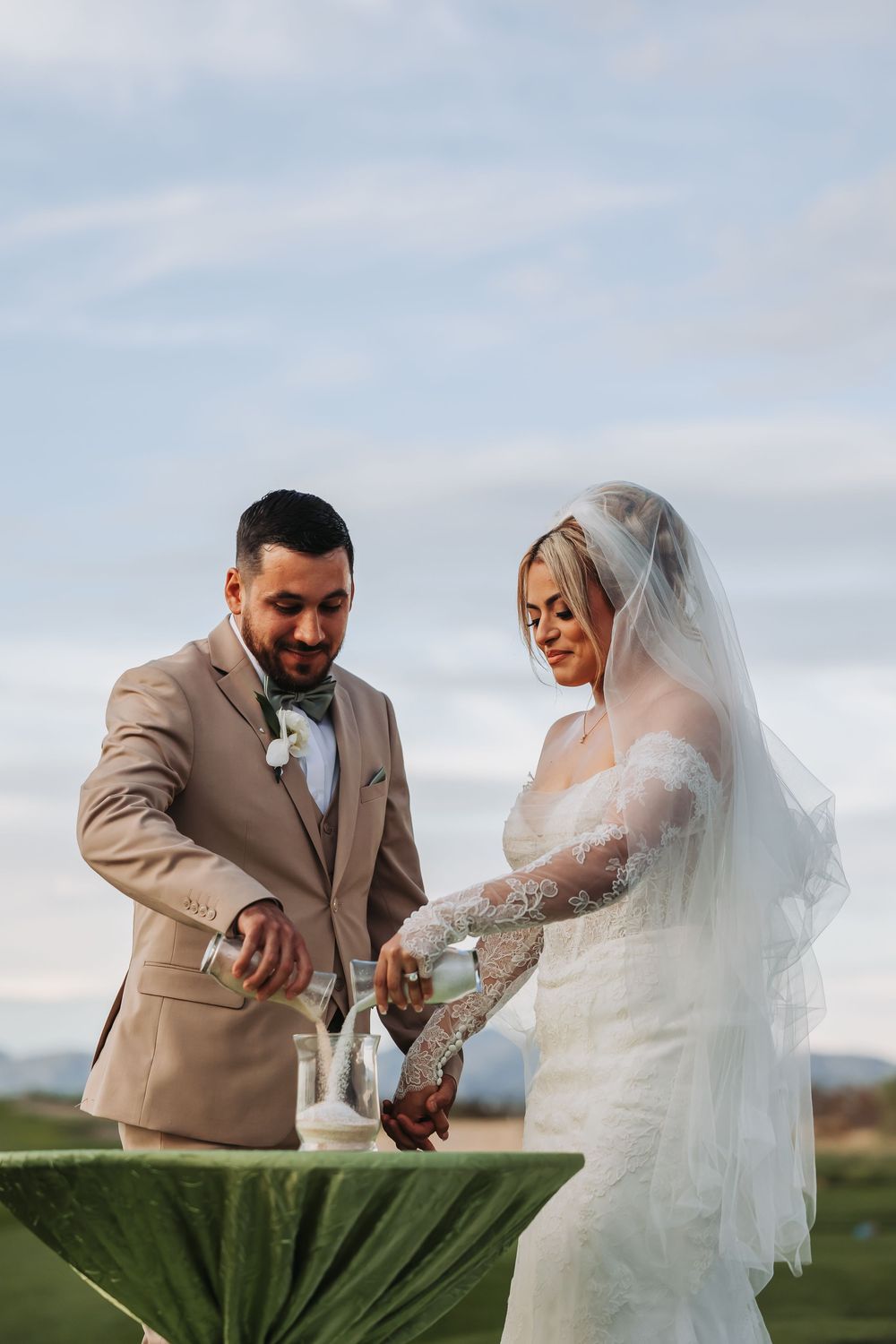 Bride and groom performing a sand unity ceremony outdoors, bride in lace gown with veil, groom in tan suit.