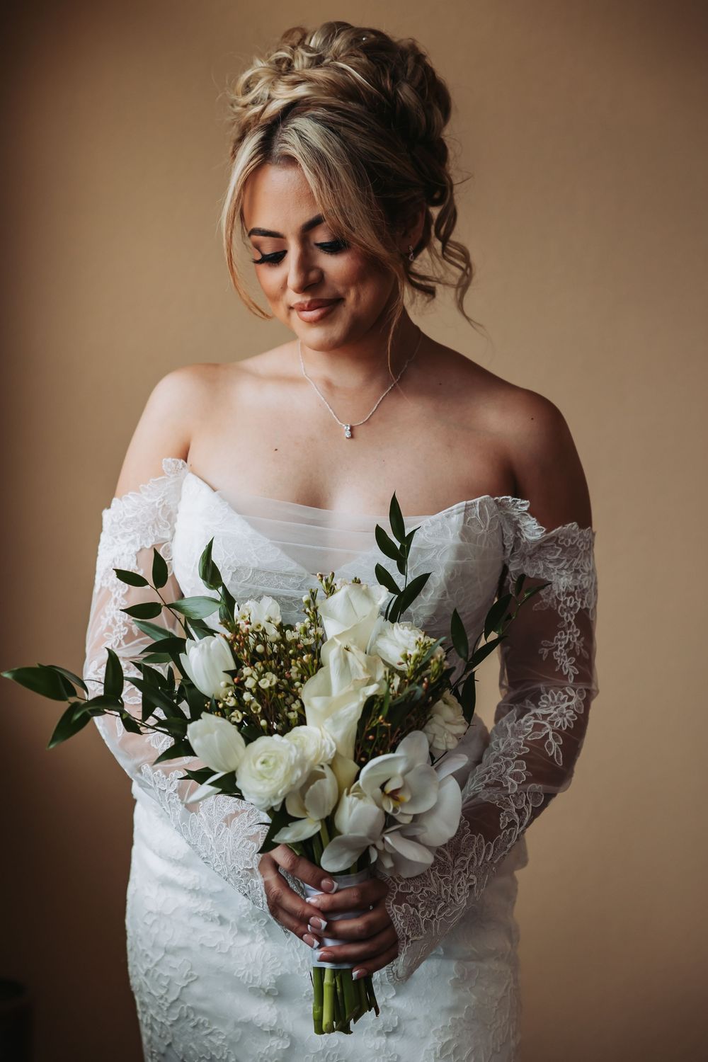 Bride in off-shoulder lace gown holds white floral bouquet with orchids and greenery, smiling downward.