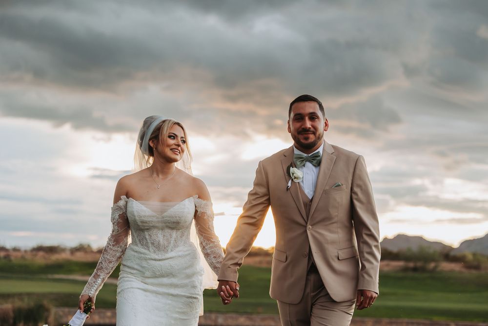Bride and groom holding hands walking outdoors under dramatic cloudy sky at sunset in wedding attire.
