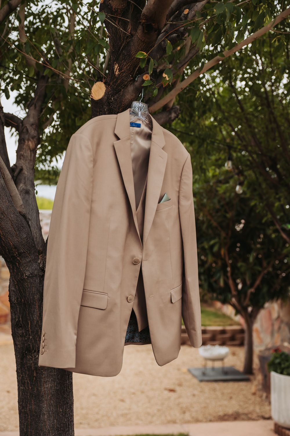 Beige men's blazer with green pocket square hanging on a tree branch outdoors at a wedding venue.