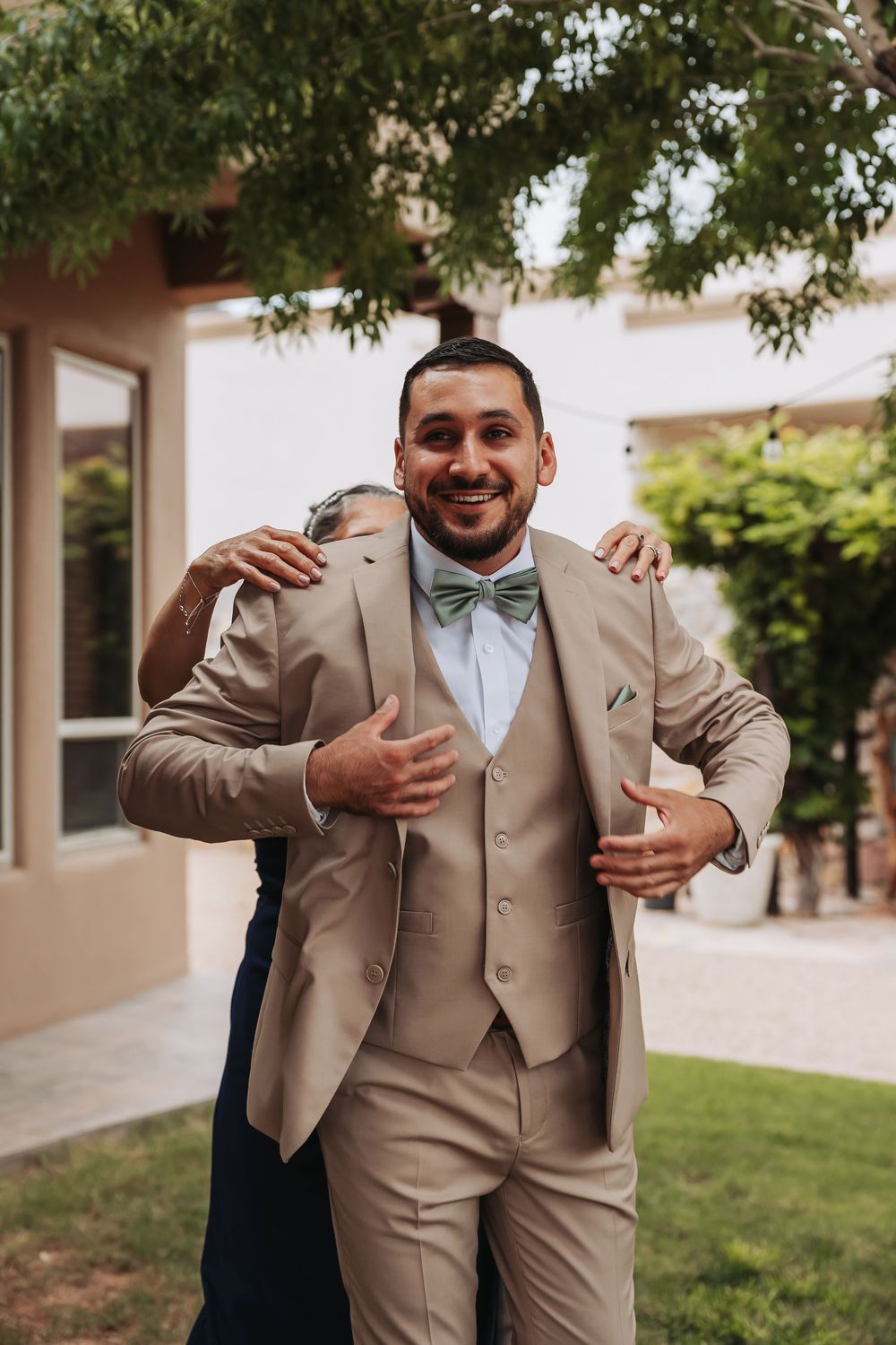 Smiling groom in tan three-piece suit with sage bow tie being hugged outdoors on wedding day.