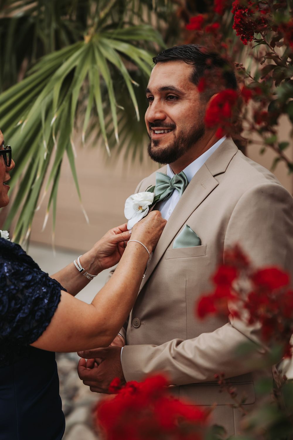 Smiling groom in tan suit with sage bow tie gets boutonniere pinned by woman, surrounded by red flowers.