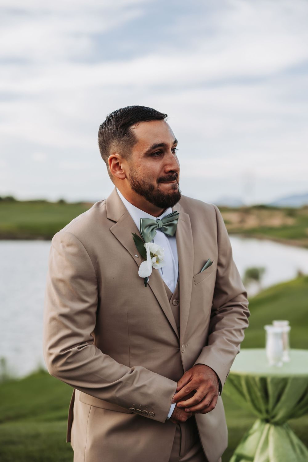 Groom in tan suit with floral boutonniere stands outdoors near a lake at a wedding ceremony.