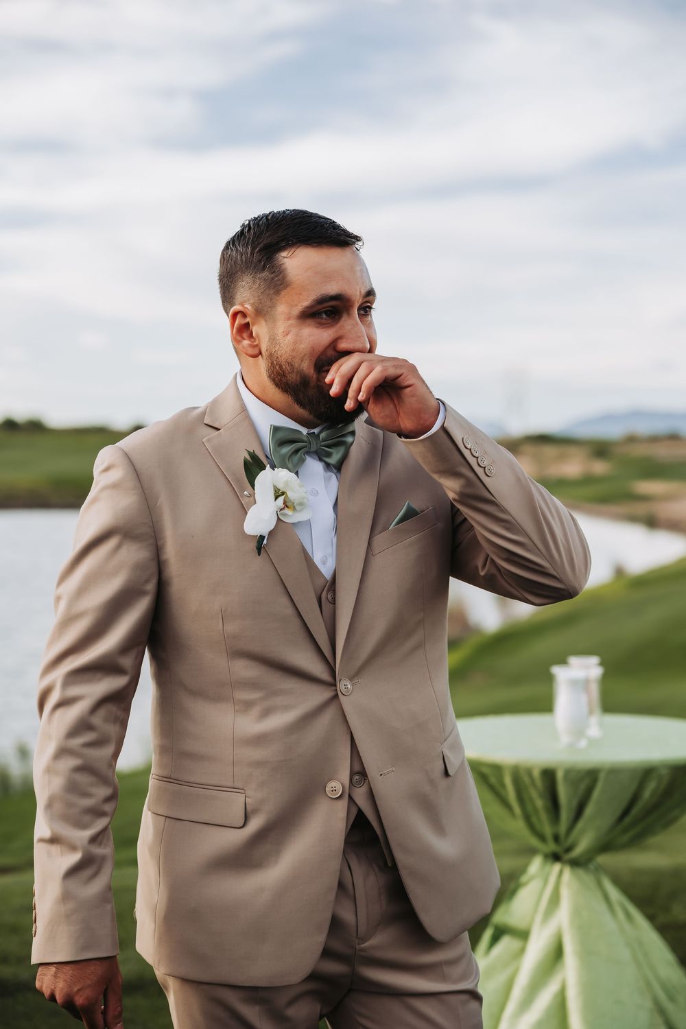 Emotional groom in tan three-piece suit with white boutonniere wiping tear at outdoor wedding ceremony.