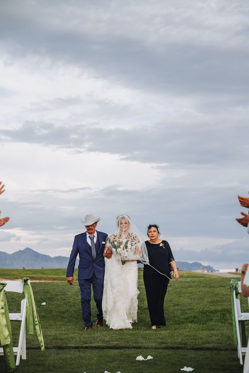 Bride walks down outdoor aisle flanked by man in cowboy hat and woman, mountains visible under cloudy sky.