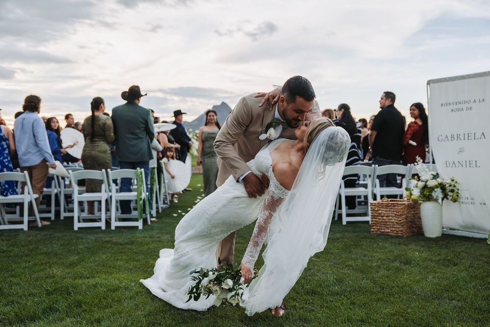 Bride and groom share a romantic dip kiss at outdoor wedding ceremony with guests seated in white chairs.
