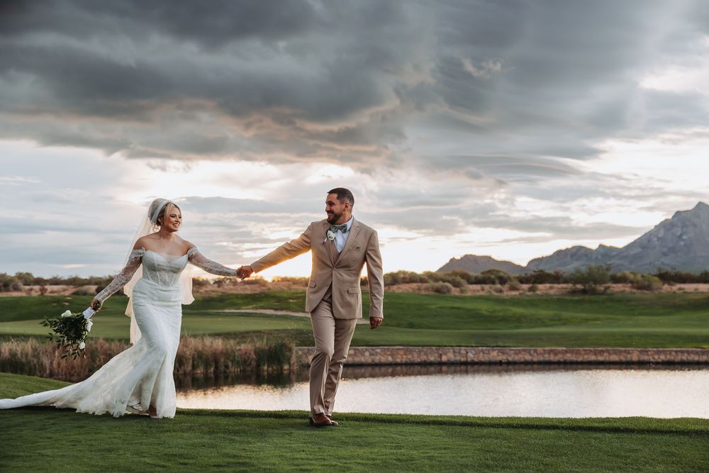 Bride and groom hold hands walking on lush golf course with mountain backdrop and dramatic sunset sky.