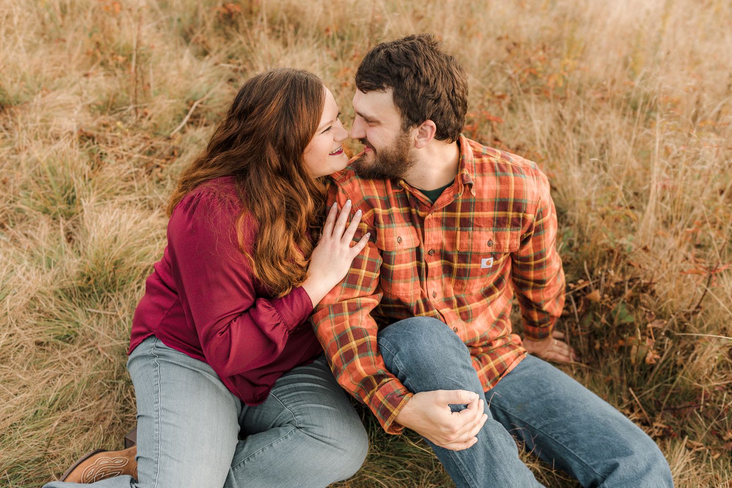 A couple sits together in a golden field, sharing a romantic moment in casual fall outfits.