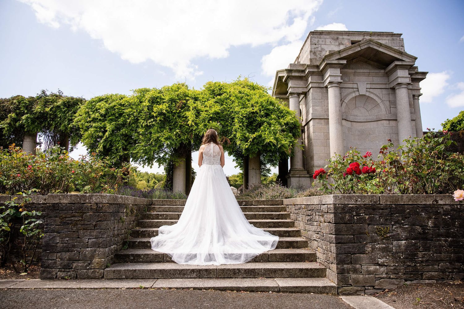 Bride in a long white wedding gown with a flowing train stands on stone steps at memorial gardens Dublin photo by AK wedding photography.