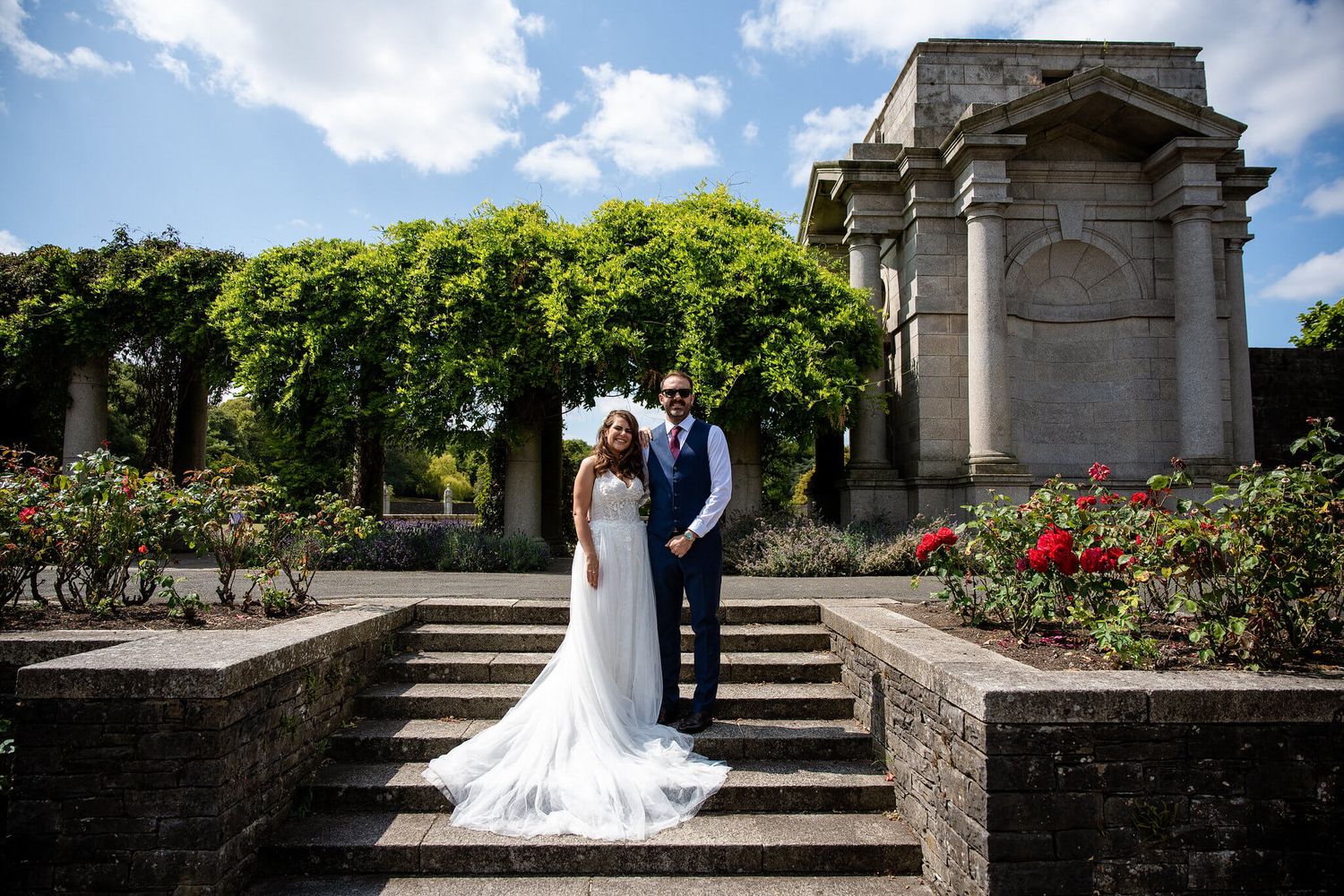 Bride in a long white gown and groom in a navy suit pose together on stone garden steps beside blooming red roses and a classical monument.