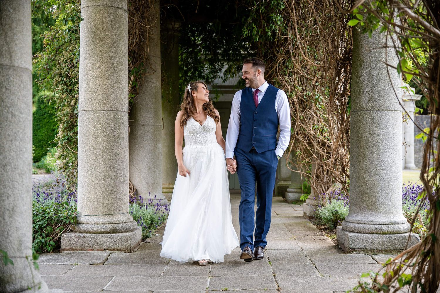 Bride in a white wedding gown and groom in a navy vest hold hands while walking through a stone colonnade photo by AK wedding photography.