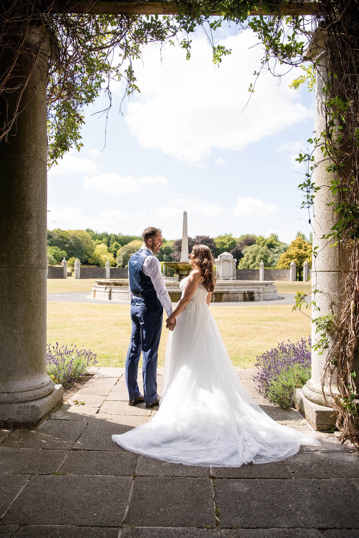 Bride in a flowing white gown holds hands with a groom in a navy suit at war memorial garden Dublin photo by AK wedding photography.