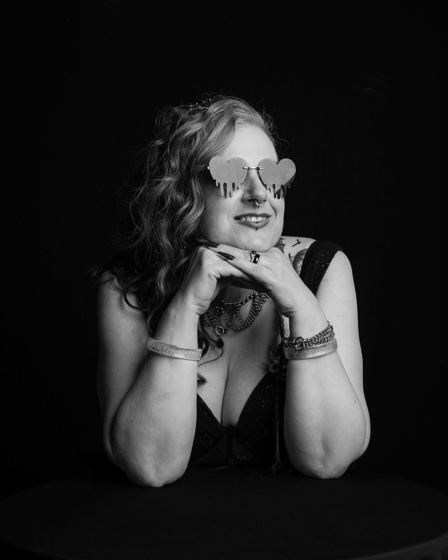 Black and white studio portrait of a smiling woman with wavy hair, heart-shaped sunglasses and layered bracelets, resting her chin on claspe