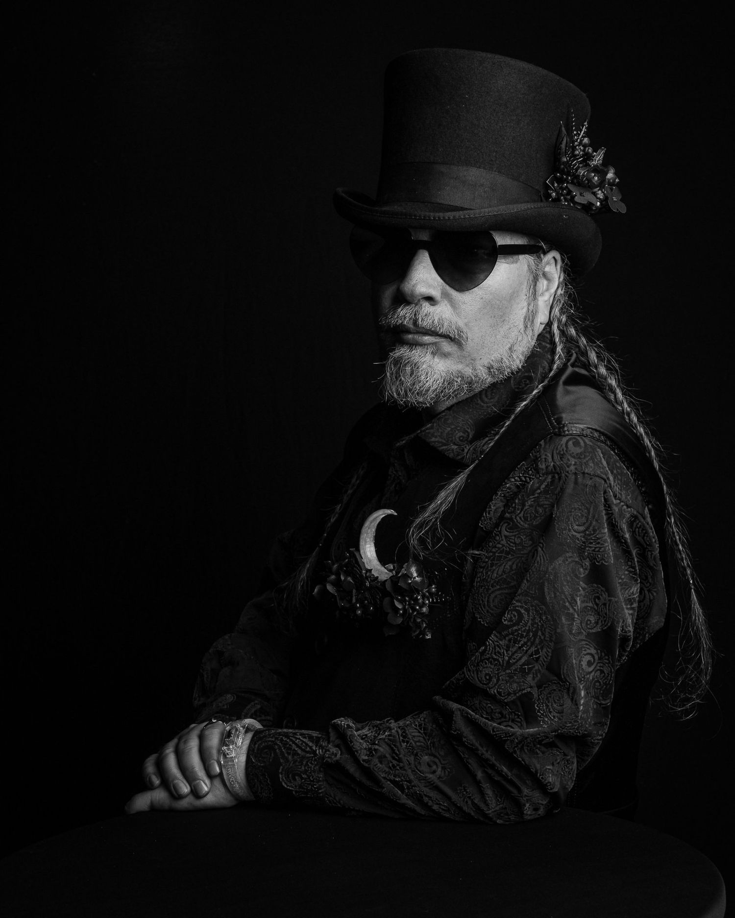 Black and white portrait of a bearded man in a top hat, heart-shaped sunglasses and a paisley shirt with a crescent moon pendant.