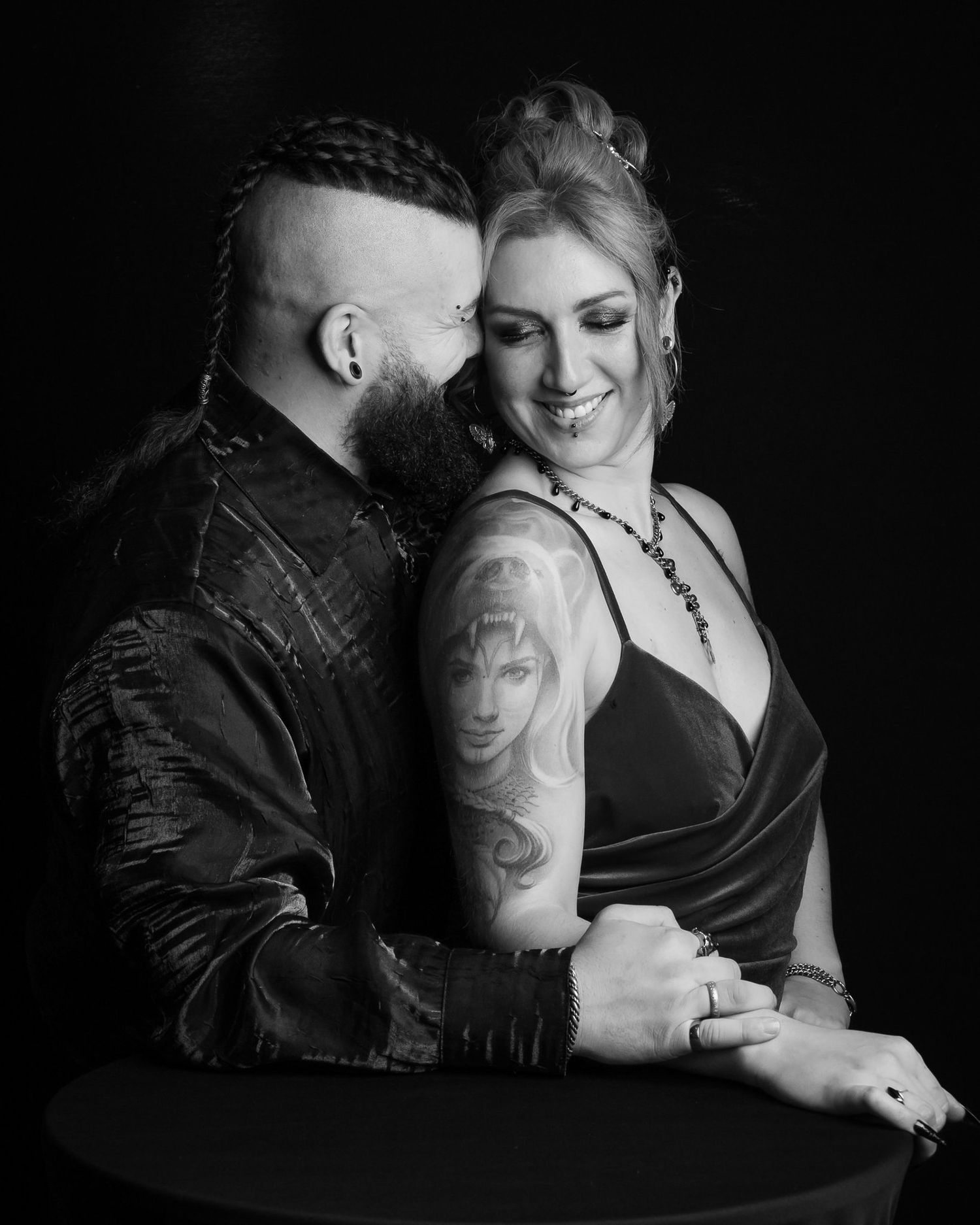Black and white portrait of a bearded man with a braided mohawk embracing a smiling woman with a portrait tattoo on her arm.