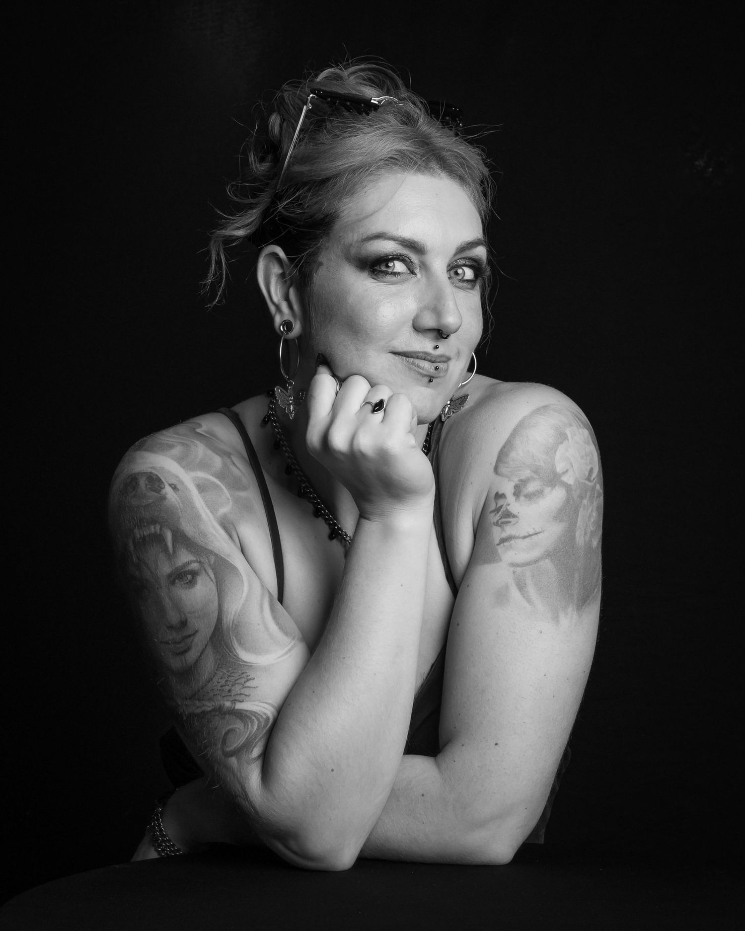 Black and white portrait of a smiling tattooed woman with updo hair and sunglasses on her head, resting her chin on her hand.