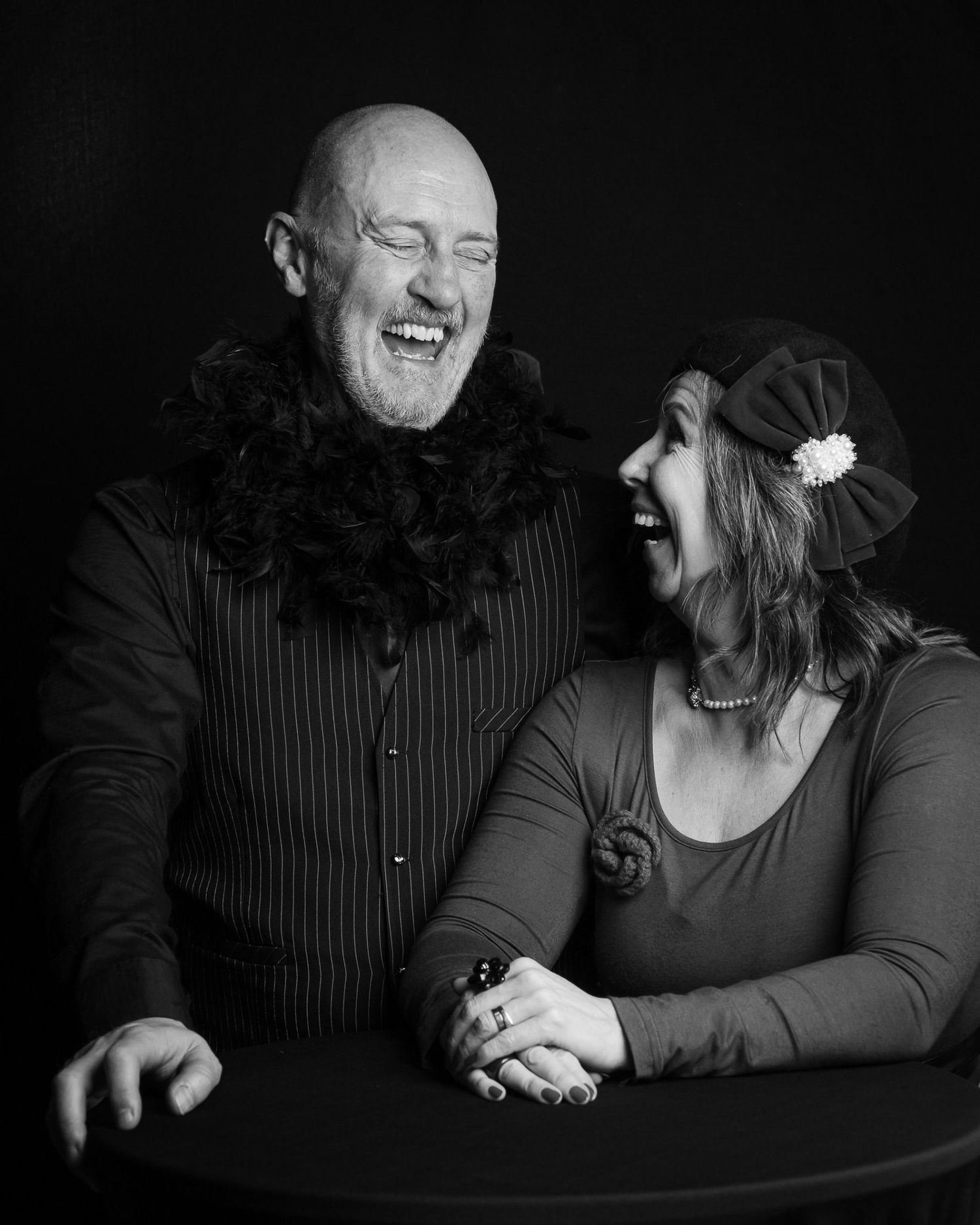 Black and white portrait of a laughing bald man in a black feather boa and a smiling woman in a flapper-style headband holding hands.