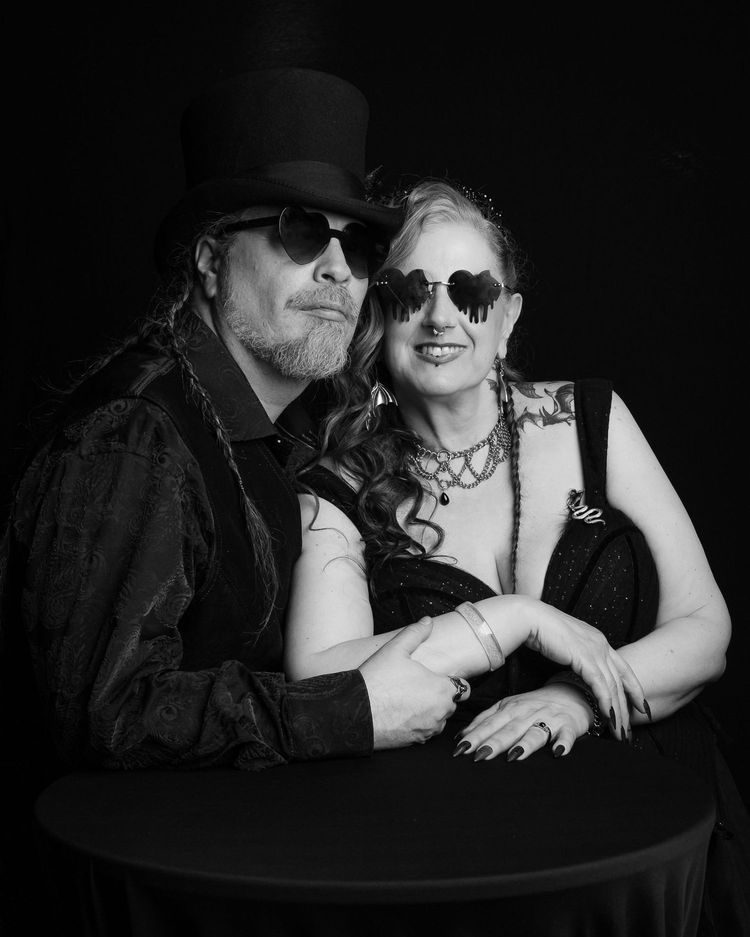 Black-and-white portrait of a couple in gothic attire, he in a top hat and sunglasses, she in heart-shaped shades, posing close at a table.